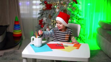 Noel Baba şapkalı küçük mutlu çocuk bir fincandan bir sürü kalem alıyor. Evde çizim yapan sevimli bir çocuk. Bulanık arkaplanda Noel ağacı.