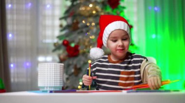 Sevimli beyaz erkek bebek masa başında oturup kalemleri karıştırıyor. Noel Baba şapkalı çocuk evde resim çiziyor. Bulanık arkaplanda Noel ağacı.