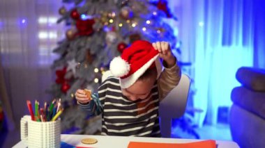 Sevimli küçük çocuk kırmızı Noel Baba şapkasını çıkarıyor. Tatlı çocuk kurabiye yiyor ve oynaşıyor. Bulanık arkaplanda Noel ağacı.