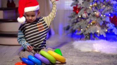 Kırmızı Noel Baba şapkalı çocuk süslü Noel ağacının yanında yerde oturuyor. Bebek renkli piramitlerle oynuyor..