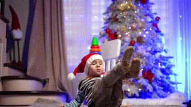 Kırmızı Noel Baba şapkalı neşeli çocuk dans ediyor ve yerde yuvarlanıyor. Mutlu bebek Noel ağacının yanında.