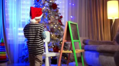 Kırmızı Noel Baba şapkalı küçük bir çocuk tahtaya gelir ve onu toz beziyle temizler. Bulanık arkaplanda Noel ağacı.