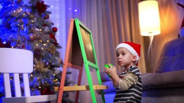 Beyaz, kırmızı şapkalı çocuk tahtayı temizliyor. Noel için dekore edilmiş küçük çocuk odada..