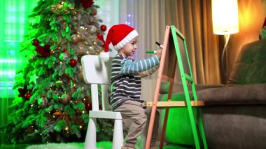 Kafkasyalı erkek çocuk tahtaya kalemlerle çizim yapıyor. Arkaplanda güzel süslenmiş Noel ağacı.