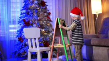 Noel Baba şapkalı genç bir çocuk tahtayı dikkatlice temizliyor. Noel için evde sevimli bir çocuk var. Arkaplanda süslenmiş köknar ağacı.