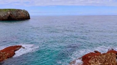 Kayalık sahildeki su yatağında kırmızı kaya oluşumları. Bulutlu gökyüzünün altında engin deniz manzarası. Suyun üstünde drone görüntüleri.