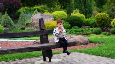 Tatlı bebek bankta oturmuş atıştırıyor. Yazın güzel bir parkta küçük bir çocuk..