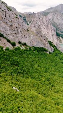 Yeşilliklerle kaplı dağın yamacında uçuyorlar. Kayaların üzerindeki hava manzarası güzel göle iniyor. Dikey video.