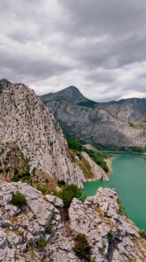 Yüksek sarp kayalar ve nadir bitki örtüsü büyüyor. Dağın yukarısındaki insansız hava aracı görüntüleri. Ayağında turkuaz bir göl var. Dikey video.