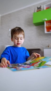 Güzel beyaz çocuk masasında oturmuş renkli bir kitabın sayfalarını çeviriyor. Çocuk resimlere bakıp yorum yapıyor. Dikey video.