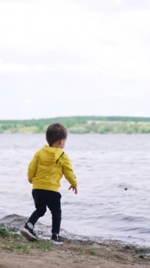 Sarı kazaklı çocuk rüzgarlı bir günde nehirde duruyor. Mutlu çocuk çakıl taşlarını suya atıyor. Dikey video.