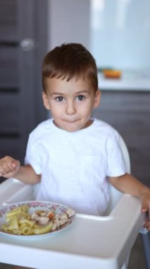 Sevimli çocuk tabağını işaret ederek yüksek sandalyede oturur. Küçük bebek evde yemek yiyor. Dikey video.