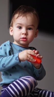Oyuncakların arasında yerde oturan bebek kameraya bakıyor. Toddler plastik bir oyuncakla oynuyor ve onu ellerinin arasına sıkıştırıyor. Düşük açı. Dikey video