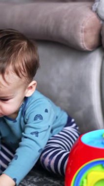 Koyu saçlı, beyaz bir çocuk kanepenin yanında yerde oturuyordu. Sevimli çocuk önünde parlak oyuncaklarla oynuyor. Dikey video