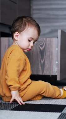 Güzel beyaz bir erkek bebek mutfak zemininde oturuyor. Çocuk kapağı konteynırdan alıp çekmeceye koymuş. Dikey video