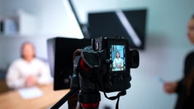 Kamera, masada oturmuş konuşan uzun saçlı bir kadının videosunu çekiyor. Stüdyoda bir blogcunun çalışması.