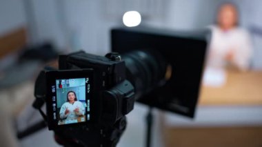 Kamera, beyaz bluzlu, uzun saçlı beyaz bir kadının masada oturup konuştuğu bir video kaydediyor. Blog konsepti. Kapatın. Bulanık arkaplan.