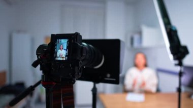 Stüdyoda kamera ve ışıklar açık. Masada oturmuş konuşan bir kadın blogcunun videosunu kaydediyorum. Bulanık arkaplan.