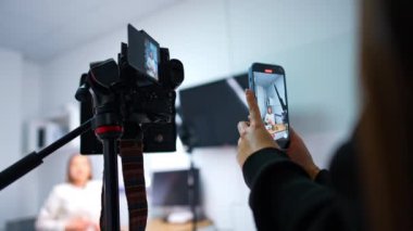 Tanımlanamayan bir kadın elinde akıllı bir telefonla video kaydediyor. Profesyonel kamera, masada oturan bir kadını çekiyor..