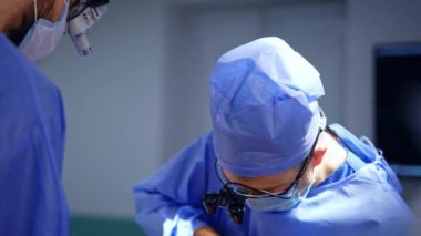 Cerrahi loupes 'taki doktor odaklanmış görünüyor. Beyin cerrahı, tıbbi bir aletle çok çaba sarf ediyor. Asistanın elinde bir kan emici var..