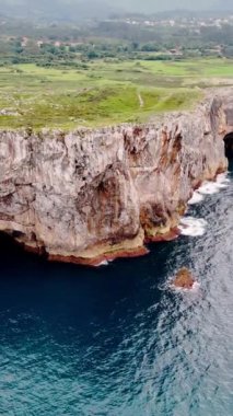 Çimlerle kaplı düz tepeli güçlü bir kaya. Mavi denizin suları kayalıklara ulaşır. Hava görüntüsü. Dikey video.