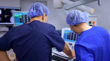 İki erkek doktor kamera karşısında sırtüstü bilgisayar ekranına bakıyor. Cerrahlar hastanın durumuyla ilgili danışmanlık yaparlar..