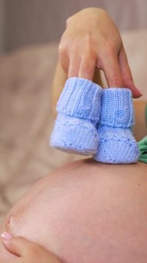 Hamile bir kadın bebek paçalarını tutuyor, yakın plan. Göbeğinde mavi ayaklı sekiz aylık hamile bir kadın. Dikey video
