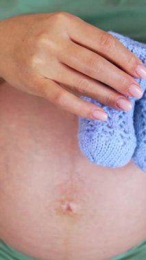 Hamile, beyaz kadın yatağında oturuyor, göbeğinde de örgü çizmeler var. Kapatın. Dikey video