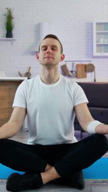 Genç adam mutfakta yoga pozunda oturuyor. Beyaz tişörtlü adam antrenmandan sonra dinlenme egzersizi yapıyor evde bir paspasın üzerinde otururken dikey video.