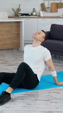 Adam içeride bir paspasın üzerinde rahatlıyor. Evdeki sıkı çalışmadan sonra paspasın üzerinde oturan genç adam. Karantina sırasında zindelik, dikey video.