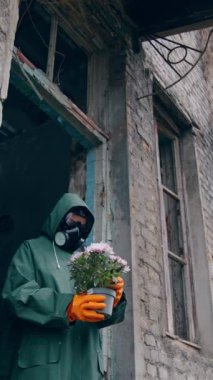 Terk edilmiş bitkilerle birlikte duran bir ekolojist. Koruyucu üniformalı ve gaz maskeli bir adam yıkılmış binanın yanında çiçek tutuyor ve ona bakıyor. Kimyasal saldırı ve hava kirliliği konsepti. Dikey video