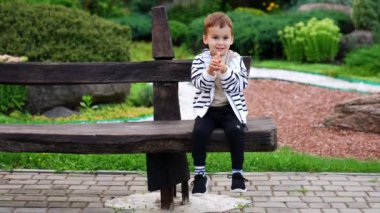 Şirin gülümseyen bebek parkta ahşap bankta oturuyor. Güzel çocuk ellerini sallıyor..