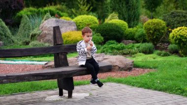 Güzel beyaz erkek bebek parktaki ahşap bankta oturuyor. Çocuk atıştırmalık bir şeyler yiyor..