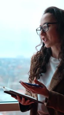 Genç, kıvırcık, uzun saçlı esmer kadın elinde kağıt bir kitapla pencerede duruyor. Soru soran ve not alan gülümseyen kadın. Portreyi kapat. Dikey video