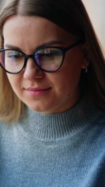 Gözlüklü sarışın kadın dizüstü bilgisayarına odaklanmış. Kadın bir şey yazıyor, sonra kameraya bakıyor ve gülümsüyor. Arkaplan bulanık. Dikey video