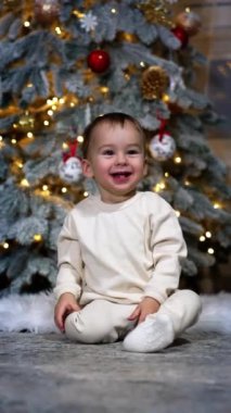 İnanılmaz sevimli küçük çocuk mutlu bir şekilde gülüyor ve kameraya doğru sürünüyor. Arka planda süslenmiş Noel ağacı. Dikey video