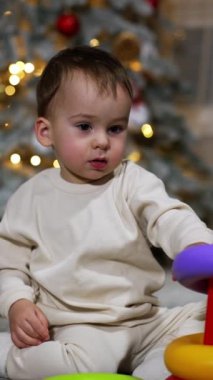 Güzel, sakin bir erkek bebek yerde oturan oyuncak bir piramitle oynuyor. Noel için dekore edilmiş bir oda. Dikey video