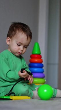 Huzurlu erkek bebek yerde oturuyor. Çocuk kordonu elinde geriyor. Oyuncak kutusu ve oyuncaklar etrafa saçılmış. Dikey video