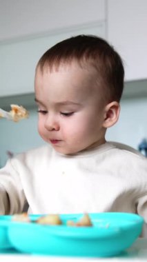 Bebek yemek yiyor ve kaşık tutuyor. Annem bir çocuğa tam kaşık veriyor. Sağlıklı beslenme. Dikey video
