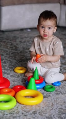 Küçük çıplak ayaklı çocuk oyuncaklarla çevrili halının üzerinde oturuyor. Beyaz çocuk piramidi birleştirirken renkleri öğreniyor. Dikey video