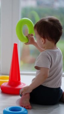 Beyaz bir çocuk kameraya doğru sırtüstü yerde oturuyor. Güzel çocuk oyuncak piramidiyle oynuyor. Dikey video