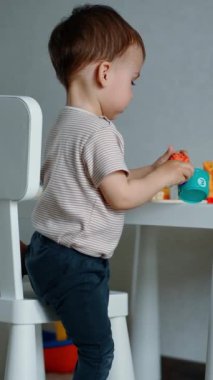 Güzel çocuk oyuncaklarıyla küçük beyaz masada oynuyor. Çocuk oyuncak kaplanı alır ve ona bakar. Dikey video