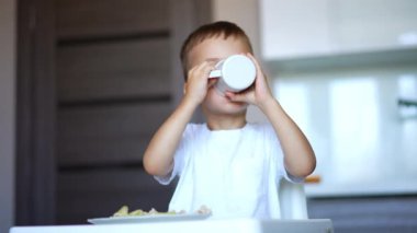 Beyaz tişörtlü çocuk bardaktan içiyor. Çocuk içkisini bitirir ve annesine bir fincan verir..