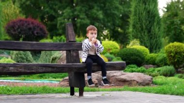 Komik çocuk parktaki bankta oturuyor. Mutlu çocuk bir oyuncak paketten atıştırıyor..