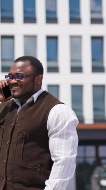 Şehirde mutlu bir Afrikalı Amerikalı adamın portresi. Havalı gözlüklü, gülümseyen siyah adam dışarıda dururken telefonla konuşuyor. Telefonu olan bir iş adamı kameraya bakar. Dikey video