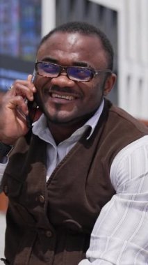 Gülümseyen afro adam dışarıda telefonla konuşuyor. Gözlüklü neşeli Afrikalı Amerikalı adam şehirde bankta oturuyor ve akıllı telefon kullanarak konuşuyor. Dikey video