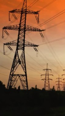 Gün batımında turuncu gökyüzüne karşı elektrik kuleleri. Akşamları yüksek voltajlı elektrik direkleri. İletim hatlarının panoramik görüntüsü. Kamera yükseliyor. Dikey video