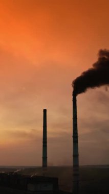 Turuncu gökyüzüne karşı endüstriyel borudan siyah duman. Gün batımında nehrin yanında siyah dumanlı fabrika bacalarının silueti. Kimyasal duman çevreyi kirletir. Ekolojik felaket. Dikey video