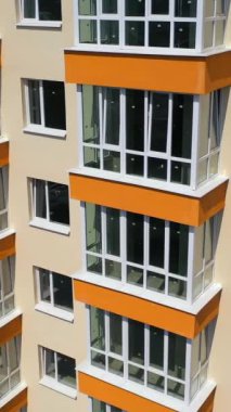Şehrin çok katlı apartmanının dış cephesi. Gökdelen binasının ön cephesinde yeni plastik pencereler ve güneş ışığında turuncu balkonlar. Dikey video