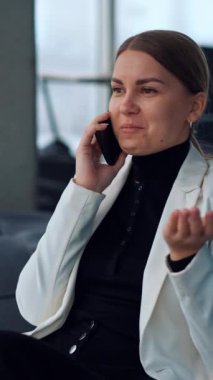 İşten izin alan ve telefonda konuşan bir kadın. Gevşemiş gülümseyen bayan koltukta oturmuş akıllı telefondan pencereye bakıyor ve sohbet ediyor. Dikey video